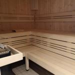 Sauna mit Holzbanken und Bodenfliesen. An der Wand hängt eine Uhr.