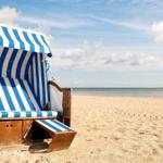 Ein blaues und weißes Strandkörbchen steht auf dem Sand. Der Himmel ist blau mit weißen Wolken.