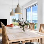 Dining room with wooden table, chairs, and sea view. Windows, TV, and flowers on table.
