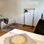Schlafzimmer mit Bett, Kleiderständer und Holzboden. Tisch mit Stuhl im Vordergrund.