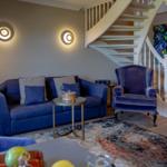 Wohnbereich mit blauem Sofa, Sessel und Treppe. Wandlampen und Teppich sind sichtbar.