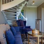 Ein Wohnzimmer mit blauem Sofa, Sessel und Spiraltreppe. Tisch und Teppich sind sichtbar.