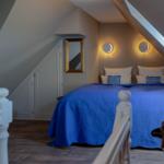 Schlafzimmer mit blauer Bettdecke, zwei runden Wandlampen und einem Spiegel an der Wand.