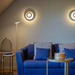 Blauer Sofa mit Kissen, runde Tische und Wandlampen in einem Raum mit schräger Decke.