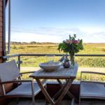 Terrasse mit Tisch, zwei Stühlen und Blumen. Blick auf grünes Feld unter blauem Himmel.