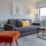 Ein Wohnzimmer mit grauem Sofa, orangefarbenem Sessel und Fensterblick auf Dächer.