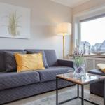 Ein Wohnzimmer mit grauem Sofa, gelben Kissen und einem Fenster mit Blick auf Dächer.