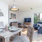 Essbereich mit Tisch und Stühlen, Wohnbereich mit Sofa und Fernseher, große Fenster mit Blick auf Garten.