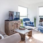 Wohnzimmer mit TV, Holztisch und blauen Sesseln. Blick auf Wasser durch Fenster.