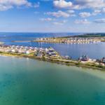 Aerial view of waterfront homes with clear water and distant horizon under blue sky with clouds.