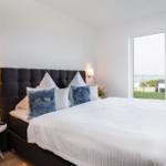 Schlafzimmer mit großem Bett, Fensterblick auf Wasser und Pflanzen auf Nachttisch.