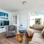 Living room with sofa, armchair, glass table, and TV. Large windows with balcony view.