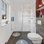 Badezimmer mit Dusche, Toilette und Fenster. Bodenfliesen und zwei runde Teppiche mit Ankern.