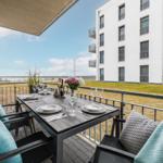 Terrasse mit Tisch, Stühlen und Blick auf Wasser und Gebäude.