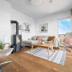 Wohnzimmer mit Kamin, Sofa, zwei Sessel und Holzboden. Blick auf Balkon mit Stadtblick.