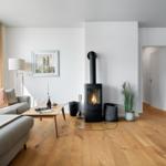 Wohnzimmer mit Holzofen, Sofa und großem Fenster. Holzboden und weiße Wände.