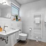 Bathroom with sink, toilet, and heater. White tiles and window with blinds.