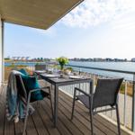 Terrasse mit Tisch und Stühlen, Blick auf Wasser und gegenüberliegende Gebäude.