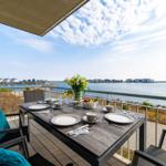 Terrasse mit Tisch, Stühlen und Meerblick. Blumen, Obst und Geschirr sind darauf platziert.