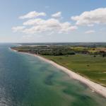Strand mit grünen Feldern und blauem Meer unter einem wolkenverzogenen Himmel.
