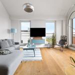 Wohnzimmer mit Sofa, Fernseher und Holzboden. Große Fenster und Pflanzen sorgen für viel Licht.