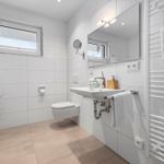 Badezimmer mit Toilette, Waschbecken und Heizkörper. Fenster, Spiegel und Pflanze sichtbar.