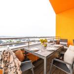 Terrasse mit Tisch, Stühlen und Sonnenblumen. Blick auf Wasser und Häuser.
