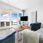 Wohnzimmer mit Blick auf Balkon und Meer. Sofa, Sessel und TV sichtbar.