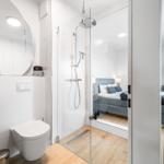 Badezimmer mit Dusche und Toilette. Im Hintergrund das Schlafzimmer mit Bett und Fernseher.