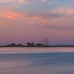 Sonnenuntergang über ruhigem Wasser mit Leuchtturm und Segelschiff im Hintergrund.