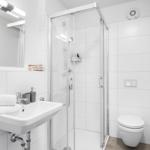 Badezimmer mit Dusche, Waschbecken und Toilette. Weiße Fliesen und bodenfarbener Fliesenboden.