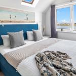 Schlafzimmer mit Blick auf Wasser und Häuser. Blauer Kopfteil, Fenster und Wandbild.
