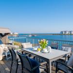 Terrasse mit Esstisch, Liegestuhl und Meerblick. Hintergrund: Wasser und Gebäude.