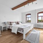 Schlafzimmer mit weißem Bett, Holzboden und zwei Fenstern mit Blick nach draußen.