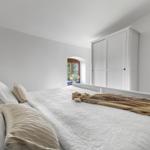 Schlafzimmer mit weißem Bett, Kleiderschrank und Fenster mit Blick nach draußen.