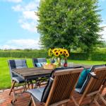 Holztisch mit Stühlen auf Terrasse. Sonnenblumen, Obst und Getränk darauf. Grünfläche und Bäume im Hintergrund.