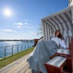 Eine Person ruht in einem Strandkorb auf einer Terrasse mit Blick auf Wasser und Himmel.