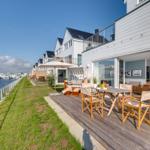 Terrasse mit Tisch, Stühlen und Sonnenschirm neben Wasser. Modernes Haus mit Glasfront.