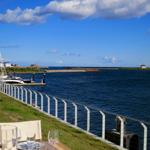 Tisch mit Besteck und Gläsern auf Grasfläche am Wasser mit Blick auf Hafen und blaues Meer.