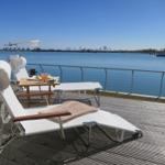 Terrasse mit Liegestühlen und Tisch am Wasser. Boot fährt im Hintergrund.