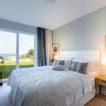 Schlafzimmer mit Blick auf den Strand und grünen Rasen. Bett, Nachttisch und Fenster sind sichtbar.