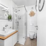 Badezimmer mit Dusche, Waschbecken und Toilette. Holzoberfläche und runder Spiegel.