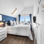 Schlafzimmer mit Blick auf Wasser, weißes Bett, blaue Wand und Dachschrägen.