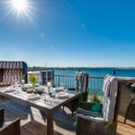 Terrasse mit Tisch, Stühlen und Grill. Blick auf Wasser und Himmel.