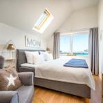 Schlafzimmer mit Dachschräge, Bett, Sessel und Fensterblick auf Wasser.