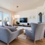 Wohnzimmer mit grauem Sofa, Fernseher und Holzboden. Große Fenster und Wandkamin.