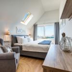 Schlafzimmer mit Dachschrägen, Bett, Sessel und Blick auf Wasser.