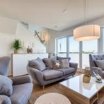 Wohnzimmer mit grauen Sesseln, Couch und Glas-Tisch. Große Fenster mit Meerblick.