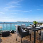 Terrasse mit Esstisch, Stühlen und Meerblick. Holzdeck, Geländer und gelbe Wand sichtbar.