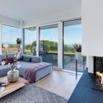 Ein Wohnzimmer mit Sofa, Kamin und großflächigen Fenstern mit Blick auf den Garten.